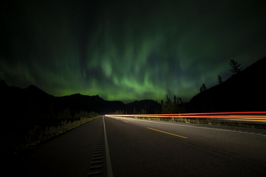 View of ethereal green aurora borealis dances above a dark highway with streaks of red taillights, framed by silhouetted mountains, Jasper, Alberta, Canada.