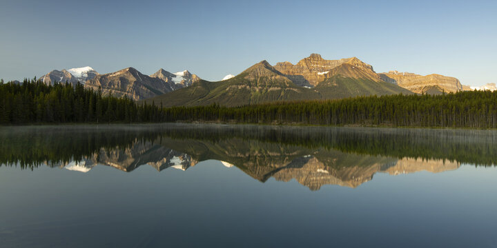 View of golden light kissing the rugged mountain peaks reflected in the still, glassy lake, framed by a dense evergreen forest, Jasper, Alberta, Canada.