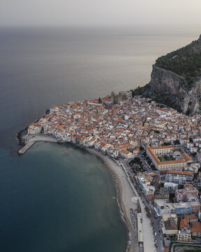 Aerial view of terracotta roofs cascading towards the azure sea, cradled by rugged cliffs in a timeless coastal embrace, Cefalu, Sicily, Italy.
