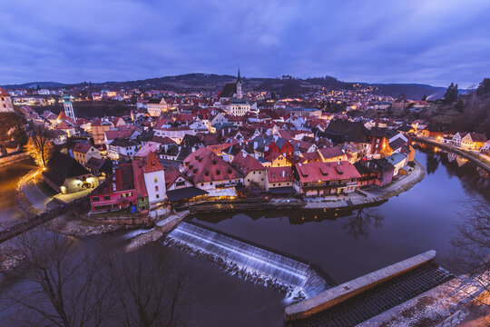 View of the Vltava River curving around the illuminated red-roofed buildings under a twilight sky, reflecting the city's charm at dusk, Cesky Krumlov, South Bohemian Region, Czechia.