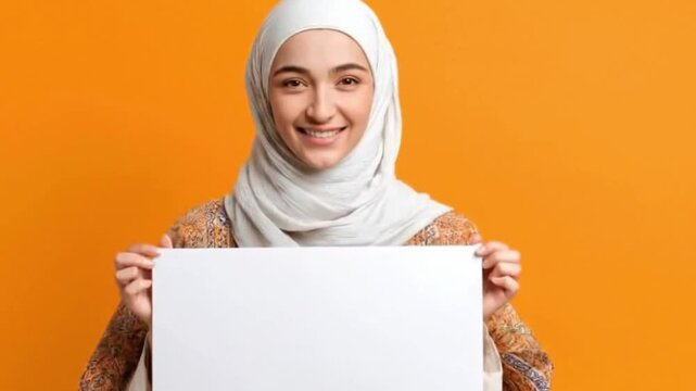A radiant young woman, adorned in a light-colored headscarf and a patterned top, smiles warmly and looks directly at the camera against a vibrant orange studio background. She holds up a pristine, bla