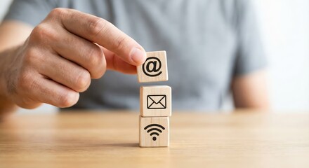 Close up of an adult man indoors stacking wooden blocks with communication icons like email and wifi on a desk, representing digital marketing and connectivity concept, soft bright morning light.