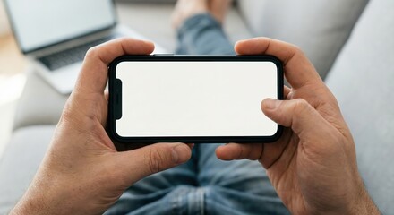 Adult man holding a smartphone with a blank white screen mockup in a horizontal orientation while relaxing indoors on a grey sofa with a laptop in a home office, technology and lifestyle concept.