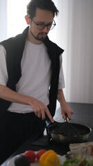 Young slim brunette man cooking scrambled eggs for breakfast in his modern stylish kitchen early in the morning