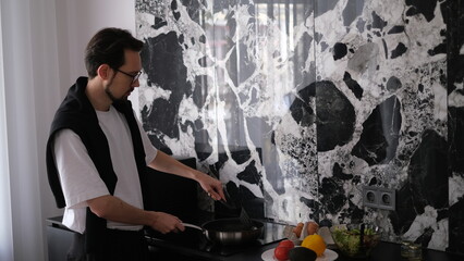 Young slim brunette man cooking scrambled eggs for breakfast in his modern stylish kitchen early in the morning