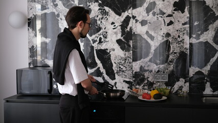 Young slim brunette man cooking scrambled eggs for breakfast in his modern stylish kitchen early in the morning