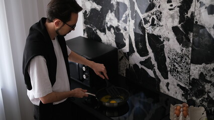 Young slim brunette man cooking scrambled eggs for breakfast in his modern stylish kitchen early in the morning