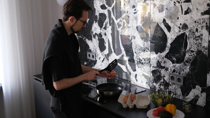 Young slim brunette man cooking scrambled eggs for breakfast in his modern stylish kitchen early in the morning