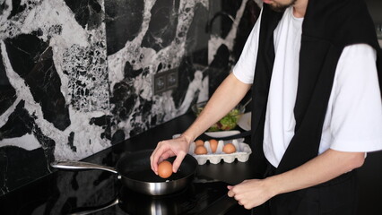Young slim brunette man cooking scrambled eggs for breakfast in his modern stylish kitchen early in the morning