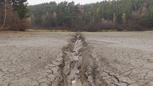 Drone footage reveals a dried lakebed.  Cracked earth patterns extend toward a forested backdrop.  A narrow stream flows through the parched landscape.  Autumn foliage colors the trees