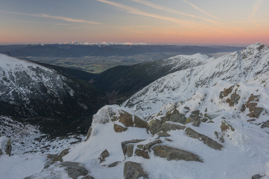 View of snow-dusted rocky peaks casting shadows over a serene valley, with a faint pink and orange sky, Zapadne Tatry, Zilina Region, Slovakia.