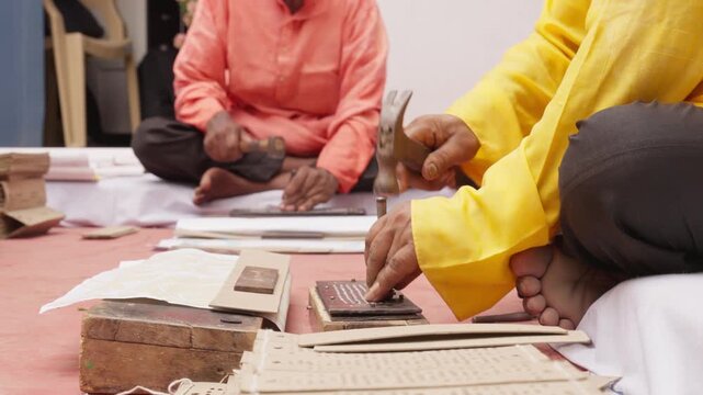 Two artisans sit and make Jacquard punch cards by hand using hammers and punches. These cards guide looms to weave detailed silk saree patterns in traditional Indian textiles like Banarasi saree