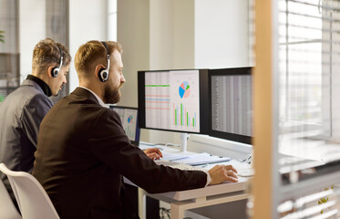 Analysts with headset support customer at office. Two men manage calls while reviewing data on screens, tracking performance and ensuring quality service for customers. Fast aid for each call.