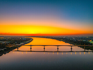 An aerial view of a city bridge spanning a wide river during sunset with soft golden light and colorful sky reflected on the water. The image conveys calm, travel, and urban scenery from day to night