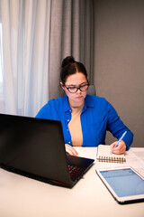 Concentrated woman with glasses types on laptop at home desk. Natural light from window highlights her productive remote work session. Focused young woman working remotely on laptop at home office