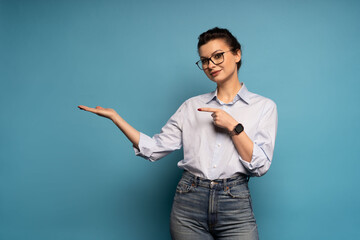 Happy business woman wearing blue shirt looking at camera pointing finger, presenting advertising offer standing isolated on blue