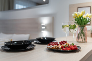 table setting in close-up.Black round plates, glasses with lemonade, raspberry tarts, a pink vase with yellow tulips. There's a bed in the background. romantic setting for a couple