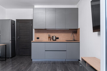 studio apartment design in modern minimalist style with built-in furniture and household appliances. small grey built-in kitchen with upper, lower cabinets, wooden countertops, black faucets, sink