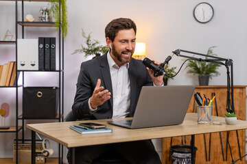 Middle-aged businessman at home office records vlog on laptop using studio microphone speaking jokes. Freelancer guy at table gestures lively smiling while sharing tips and humor for online audience.