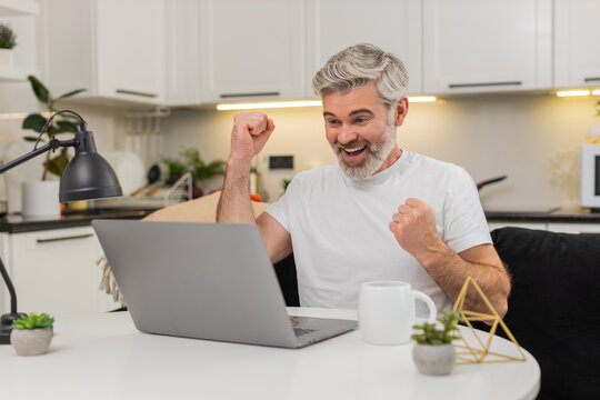 Middle-aged man at home uses laptop sees discount and promo code success smiles. Mature guy sitting at table on sofa cheers small win celebrates online game result lottery casino jackpot news smiles - Powered by Adobe