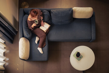 Overhead view of a young woman reading on a blue sofa with a coffee mug, home lifestyle scene with phone and tablet on a side table.