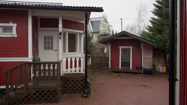 Traditional red wooden cottage and small storage shed stand in gravel courtyard. Firewood is stacked under shelter during rainy and windy autumn day in Scandinavian garden.