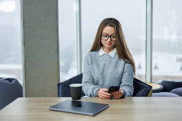 Woman using smartphone while sitting at a coffee shop table with a laptop and cup during winter season in a city location