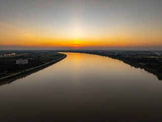An aerial view of a city bridge spanning a wide river during sunset with soft golden light and colorful sky reflected on the water. The image conveys calm, travel, and urban scenery from day to night