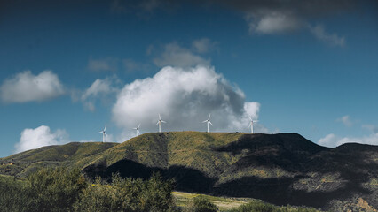 Fototapeta premium Mountain Ridge In Sicily With Wind Turbines: Green Forested Hills, Renewable Energy Structures, And Low Clouds Creating Dramatic Natural And Industrial Landscape