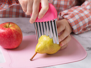 A child slicing a pear with a wavy stainless steel knife on a pink cutting board, close-up