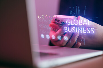 Person holding phone in front of laptop with global business text and digital graphics in a modern workspace setting