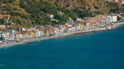 Aerial view of a vibrant coastal town nestled between a lush green hillside and the deep blue sea. It is the Giardini Naxos waterfront, located in province of Messina, Sicily, Italy. Summer morning.