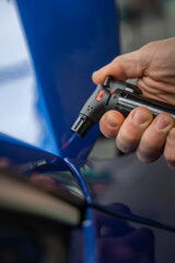 A technician uses a small butane torch to heat a tight seam on a glossy blue car panel in a dealership detailing bay, under controlled workshop lighting.