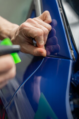 Obraz premium Hands apply and trim vinyl wrap along a blue car door edge in an indoor dealer workshop, using a precision tool and a green squeegee near the side mirror.