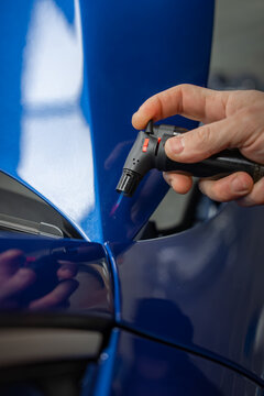 A technician uses a small heat gun on a glossy blue car hood near a panel gap in a dealer workshop. Soft indoor lighting shows precise paint correction work.