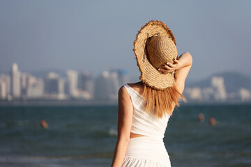 Young slim woman in wicker sun hat standing on a beach on sea waves and city background. Summer travel and vacation