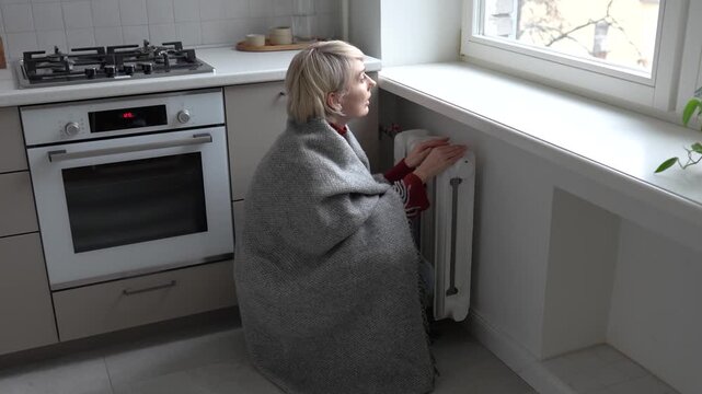 Middle aged woman wrapped in woolen plaid, sitting near window in cold unheated apartment trying to warm up hands on central heating radiator. Winter season utility problems and keeping warm indoors.