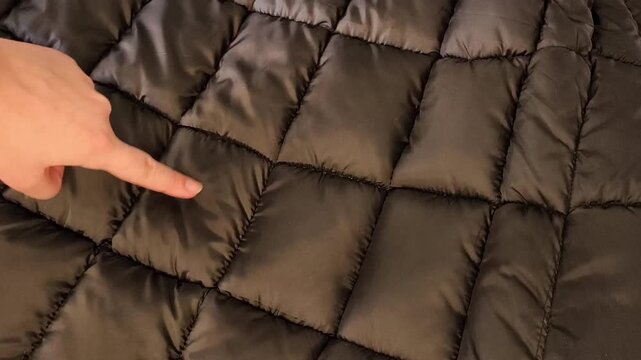 A woman presses her index finger on a quilted fabric for a down jacket. Black lining with polyester for sewing warm clothes.
