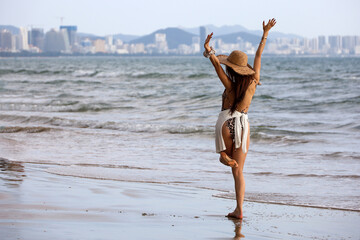 Slim happy girl in wicker sun hat standing on a beach on sea waves and city background. Happiness, freedom,  travel and vacation concept