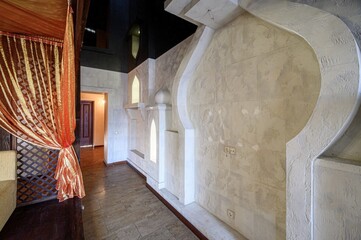 An Arabic-style interior with textured beige plaster walls, horseshoe arches, recessed niches, dark wooden floor, and rich orange-gold drapery — exotic, luxurious ambiance