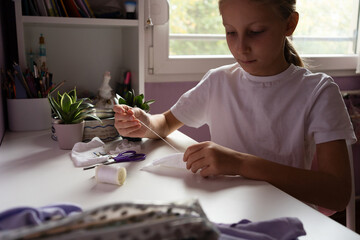 A girl sits at a white table sewing white fabric with a needle and thread surrounded by scissors and spools in a bright room with plants and bookshelf crafting hobby diy concept