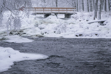 Snow blankets the ground and river in a winter scene. A wooden bridge spans the icy water, while trees are coated in frost. The water flows under the bridge