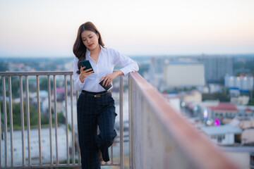 Young businesswoman standing on an urban rooftop with a city view, using her smartphone and holding a coffee cup, representing remote work, communication, and modern lifestyle