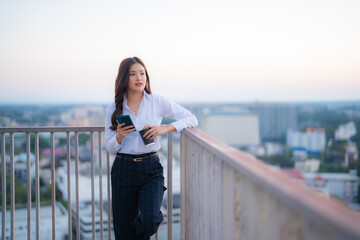Young Asian businesswoman relaxes on a city rooftop after work, holding a smartphone and coffee while gazing at the urban skyline, embodying modern professional lifestyle and work–life balance