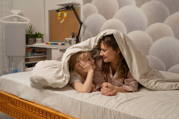 Mother and daughter hiding under a blanket in bed, sharing a secret smile. Playful moment, family bonding, cozy hideaway.