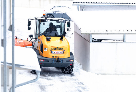 Villach, Austria - January 10, 2026: Liebherr snow plow clearing snow from a driveway in winter, showcasing heavy machinery in action with snow-covered surroundings and bright yellow color