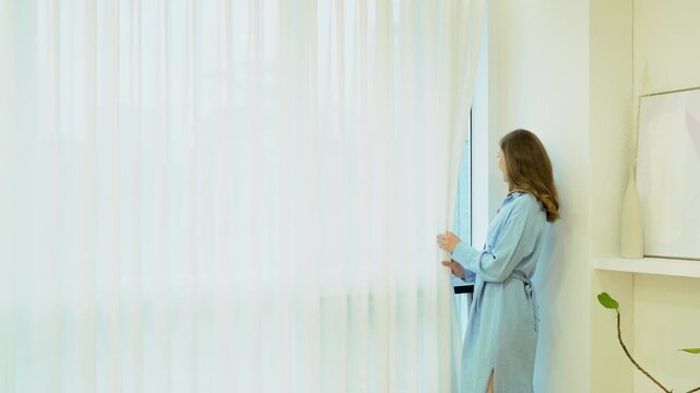Woman in blue dress stands by window, carefully pushing aside transparent curtains; light from the window highlights blue dress . Her casual pose complements calming effect of blue dress.
