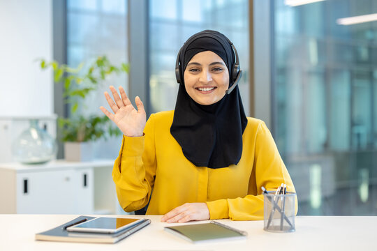 Happy muslim woman in hijab wearing a headset and waving at camera, sitting at office desk and having a video conference or providing customer support - Powered by Adobe