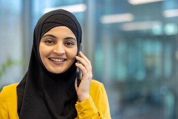 Smiling young muslim businesswoman in a black hijab and yellow blouse, confidently talking on a...