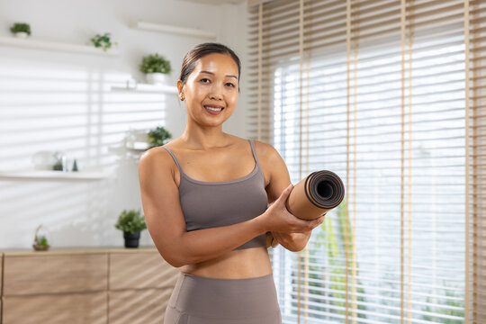 Portrait smiling asian woman holding rolled yoga mat exercise at home living room feeling happiness confident workout - Powered by Adobe